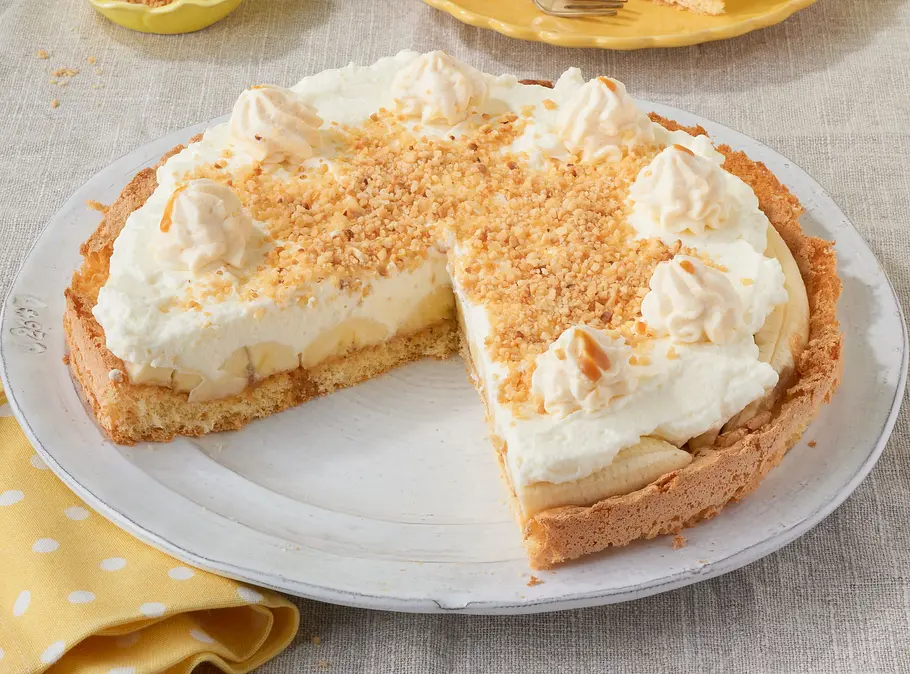 Kuchen mit knusprigem Mürbeteigboden, reichlich Sahnecreme, Bananenscheiben und Toffee, dekoriert mit Sahnetuffs und Krokantsplittern; serviert auf einem weißen Teller auf heller Stoffdecke, ein Stück auf einem gelben Teller daneben.