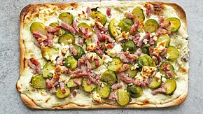 Rechteckiger Flammkuchen auf hellgrauer Steinplatte, belegt mit halbierten Rosenkohlröschen, knusprigen Baconstrreifen und zerbröseltem Käse. - Foto: Simone Neufing/MOE‘S QUICK & EASY FOOD