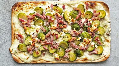 Rechteckiger Flammkuchen auf hellgrauer Steinplatte, belegt mit halbierten Rosenkohlröschen, knusprigen Baconstrreifen und zerbröseltem Käse. - Foto: Simone Neufing/MOE‘S QUICK & EASY FOOD