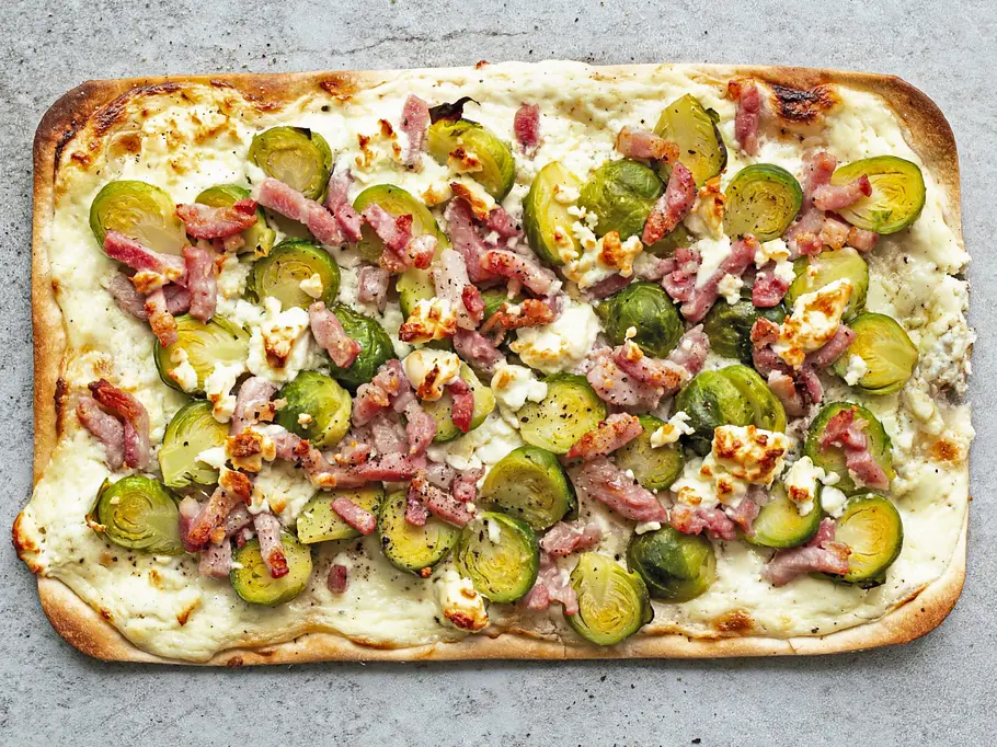 Flammkuchen mit Rosenkohl & Bacon Rezepte Rechteckiger Flammkuchen auf hellgrauer Steinplatte, belegt mit halbierten Rosenkohlröschen, knusprigen Baconstrreifen und zerbröseltem Käse.