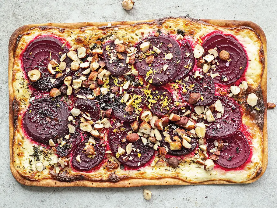 Rechteckiger Flammkuchen auf heller Steinplatte, belegt mit dünnen Scheiben Rote Bete, gehackten Nüssen, frischem Dill und Zitronenzesten.