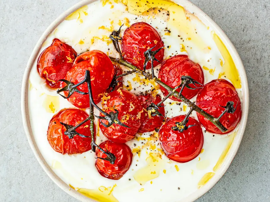 Cremig aufgeschlagener Feta serviert in einer flachen, hellen Schale, getoppt mit gerösteten Kirschtomaten am Zweig, verfeinert mit Olivenöl und Zitronenzesten.