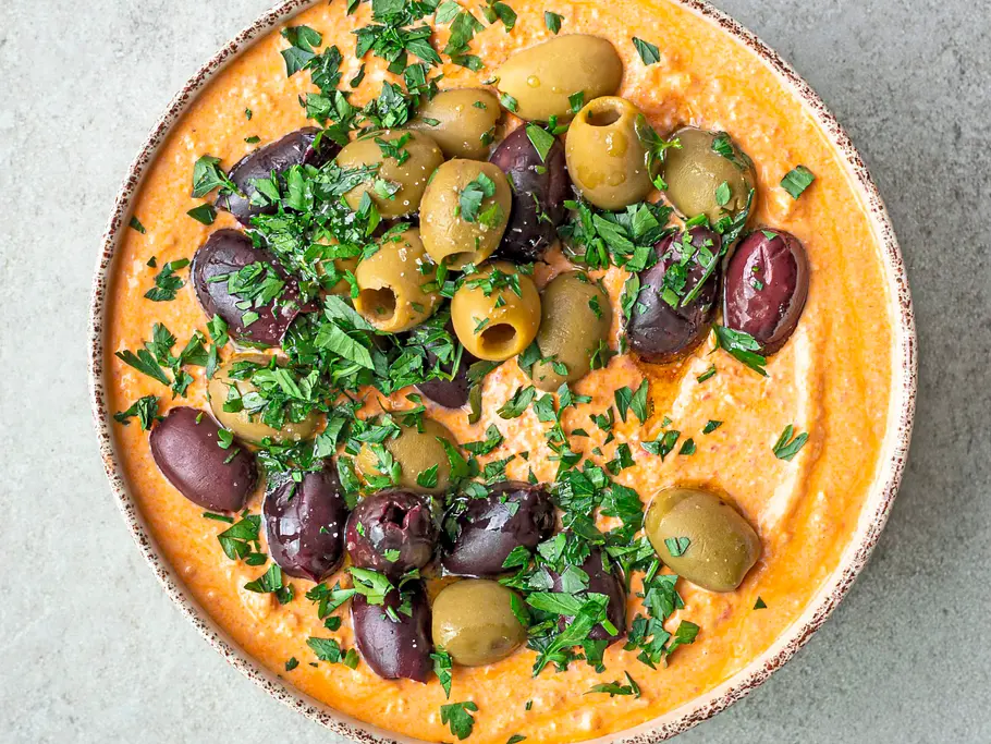 Würzig aufgeschlagener Feta mit Paprika in einer rustikalen Schale, großzügig garniert mit schwarzen und grünen Oliven sowie frischen Kräutern.