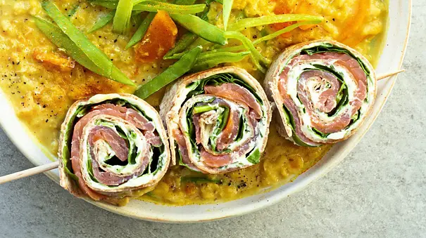 Tortilla-Röllchen mit Lachs, Frischkäse und Spinat auf einem Spieß, angerichtet auf cremiger gelben Suppe in einer weißen Schale auf heller Unterlage. - Foto: Simone Neufing/MOE‘S QUICK & EASY FOOD