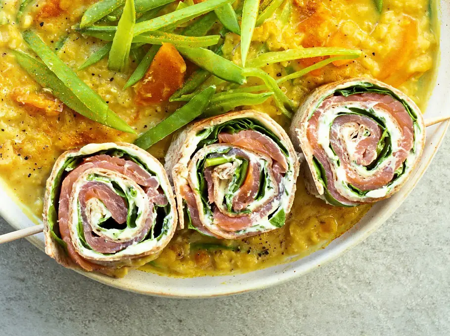 Tortilla-Röllchen mit Lachs, Frischkäse und Spinat auf einem Spieß, angerichtet auf cremiger gelben Suppe in einer weißen Schale auf heller Unterlage.