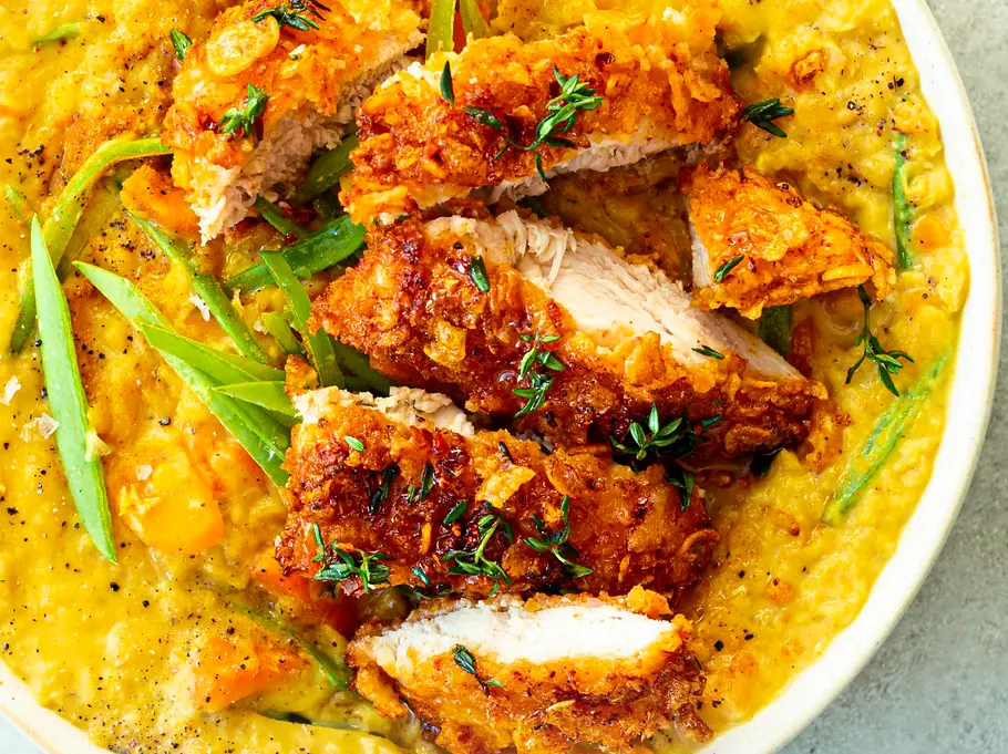 Knusprig paniertes Hähnchen, in Streifen geschnitten und mit frischen Kräutern bestreut, angerichtet auf cremiger gelben Linsensuppe in einer runden weißen Schale auf einer hellen Unterlage.