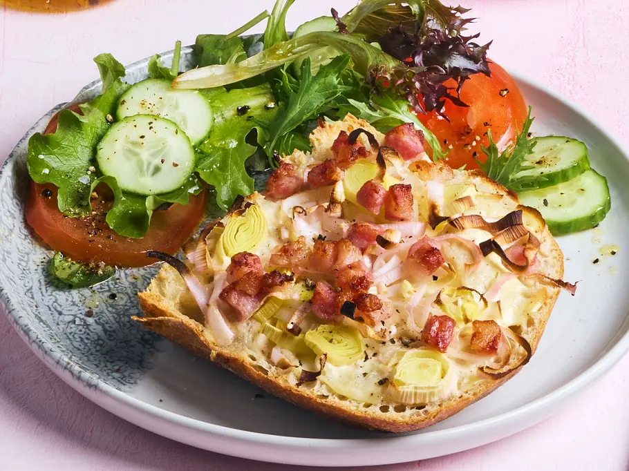 Halbierte Ciabatta-Brötchen, belegt mit Schmand, Zwiebeln, Lauch und knusprigem Speck, auf hellen Tellern angerichtet, dazu frischer grüner Salat mit Tomaten- und Gurkenscheiben. Eine kleine Kanne Vinaigrette und eine große Schüssel Salat stehen daneben auf einem zartrosa Untergrund.