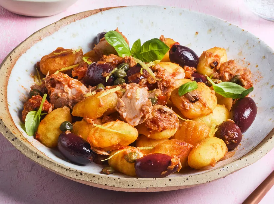 Gnocchi mit Thunfisch, schwarzen Oliven und Kapern, serviert auf einem rustikalen, weißen Teller mit Basilikumblättern als Garnitur. Daneben ein Glas Wasser und eine Kupfergabel.