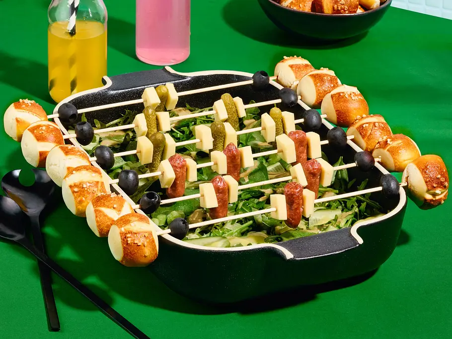 Salat-Kicker mit Laugenspießen Rezept Salat in einer Auflaufform, darauf Laugenspieße mit Mini-Laugenbrötchen, Käsewürfeln, Oliven, Würstchen und Cornichons.