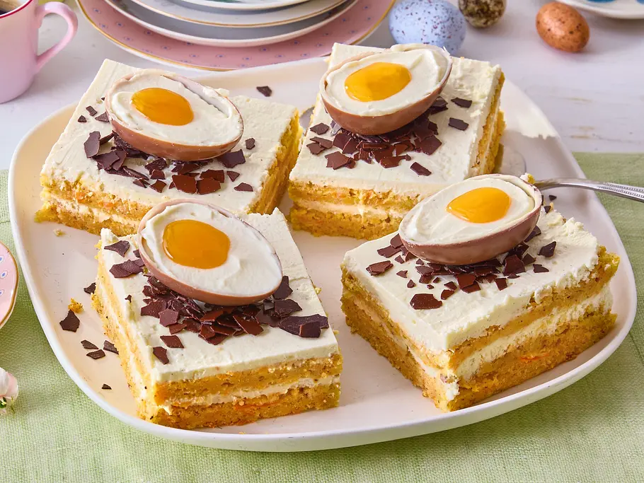 Vier rechteckige Stücke Ei-Catcher Osterkuchen mit heller Creme-Schicht und lockerem Boden, dekoriert mit Schokoraspeln und halbierten Schoko-Eiern im Spiegelei-Look, angerichtet auf einer großen weißen Platte mit grünem Tuch darunter.