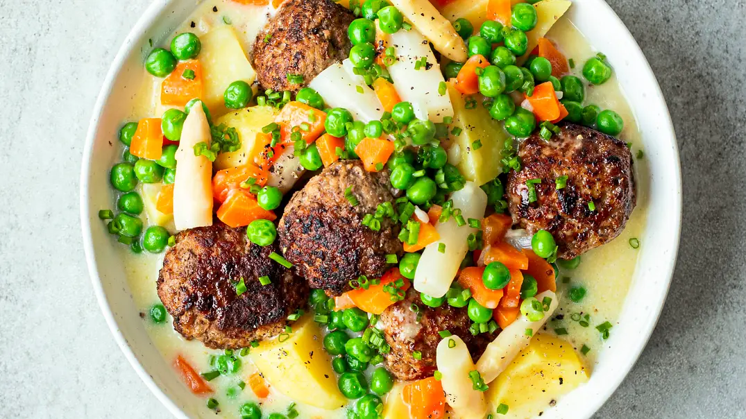 Köttbullar auf cremiger Frikasseesauce mit Kartoffeln, Erbsen, Möhren und Spargel angerichtet in einer weißen, flachen Schale. - Foto: Simone Neufing/MOE‘S QUICK & EASY FOOD