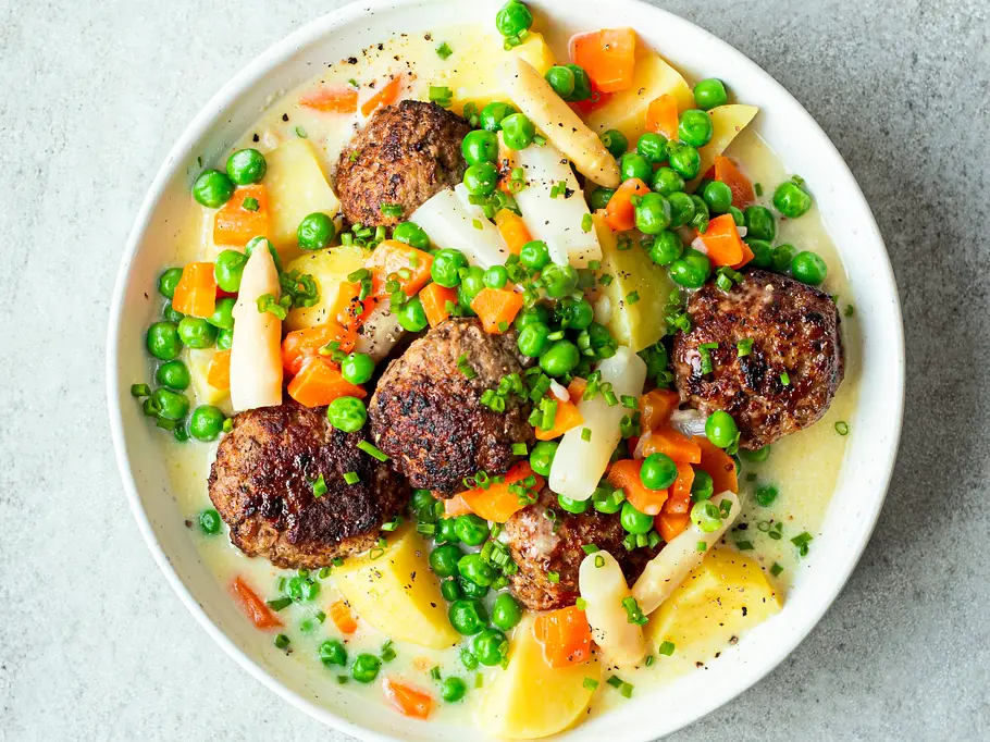 Köttbullar auf cremiger Frikasseesauce mit Kartoffeln, Erbsen, Möhren und Spargel angerichtet in einer weißen, flachen Schale.