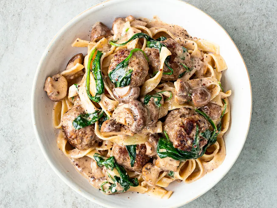 Bandnudeln mit cremiger Pilzsauce, gebratenen Köttbullar und frischem Spinat, serviert in einer weißen Schale.