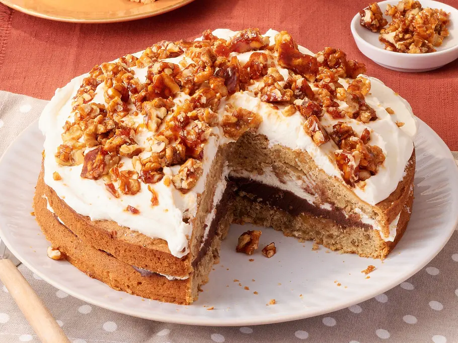 Walnuss-Espresso-Kuchen Eine angeschnittene Torte mit weißer Creme und Walnüssen steht auf dem Tisch, dahinter ein Tortenstück, Tassen und Blumen.