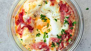 Gebackene Eier mit zartem Schinken, leicht gebräunt und mit frischen Kräutern bestreut, serviert in einer runden Glasform auf einer hellen Steinplatte. - Foto: LECKER @ Bauer Media Group