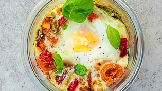 Im Glas gebackenes Ei mit goldgelbem Eigelb, umgeben von saftigen Kirschtomaten, grünem Pesto und frischen Basilikumblättern auf einer hellen Steinplatte. - Foto: LECKER @ Bauer Media Group