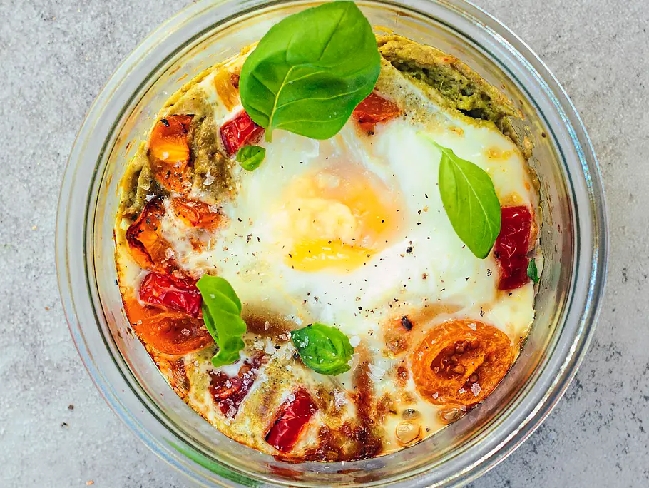 Im Glas gebackenes Ei mit goldgelbem Eigelb, umgeben von saftigen Kirschtomaten, grünem Pesto und frischen Basilikumblättern auf einer hellen Steinplatte.