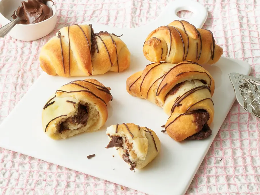 Schoko-Hörnchen aus dem Airfryer Luftige Schokoladenhörnchen mit Schokofüllung und Schokoladenstreifen auf einer weißen Platte, angerichtet auf einem rosa Tuch.