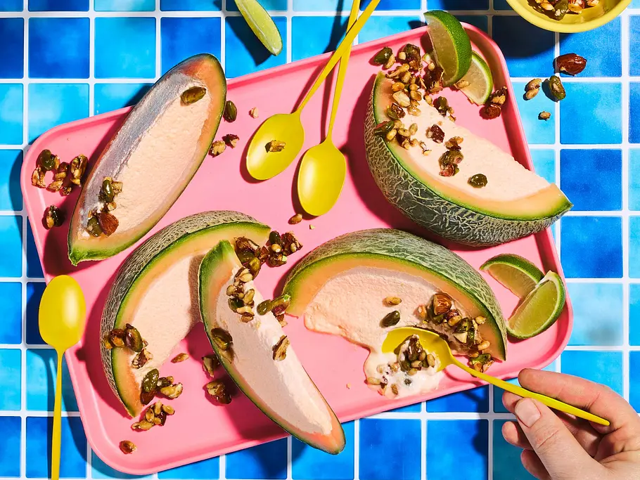 Melonen-Semifreddo in Melonenspaltenform, serviert auf einem pinken Tablett, garniert mit knusprigem Honig-Nuss-Krokant und Limettenspalten, dazu gelbe Löffel.