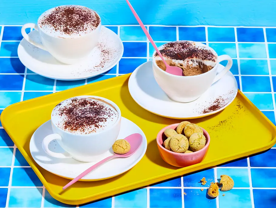 Cappuccino-Semifreddo in weißen Tassen, bestäubt mit Kakaopulver, serviert auf weißen Untertassen und einem gelben Tablett, dazu Amarettini-Kekse und rosa Löffel.