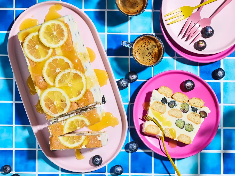 Lemon-Curd-Semifreddo à la Tiramisu Lemon-Curd-Semifreddo à la Tiramisu als rechteckige Scheibe mit Zitronenscheiben und Biskuit, serviert auf einer rosa Platte und einem pinken Teller, garniert mit Blaubeeren.