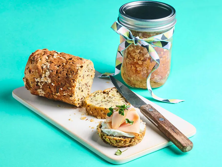 Kerniges Brot im Glas Kerniges, goldbraunes Brot auf einem hellen Schneidebrett, daneben ein angeschnittenes Brot im Glas mit Deckel und Schleife, ein Brotmesser, sowie eine Brotscheibe mit Frischkäse, Schinken und Kresse.