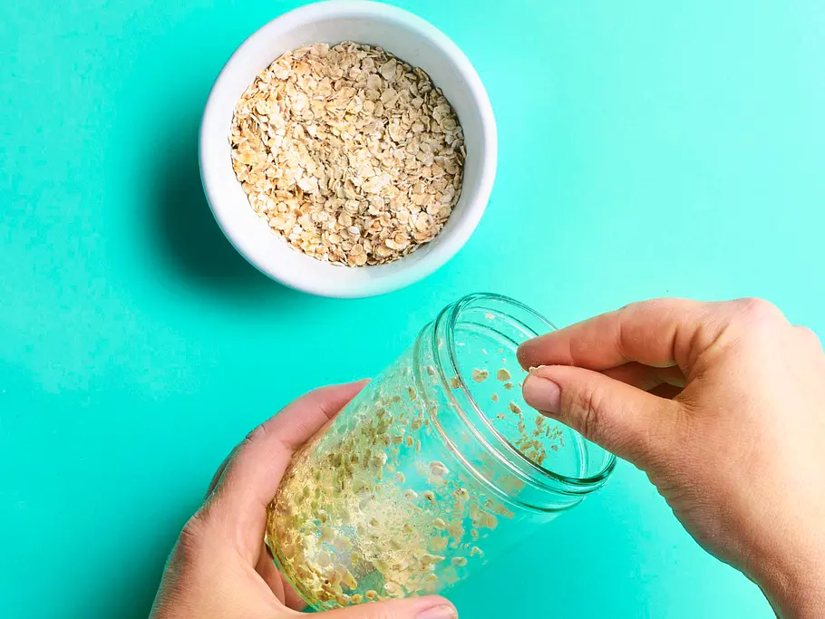 Kerniges Brot im Glas Zwei Hände fetten ein leeres Glas ein und streuen zarte Haferflocken hinein, daneben steht eine kleine Schale Haferflocken.