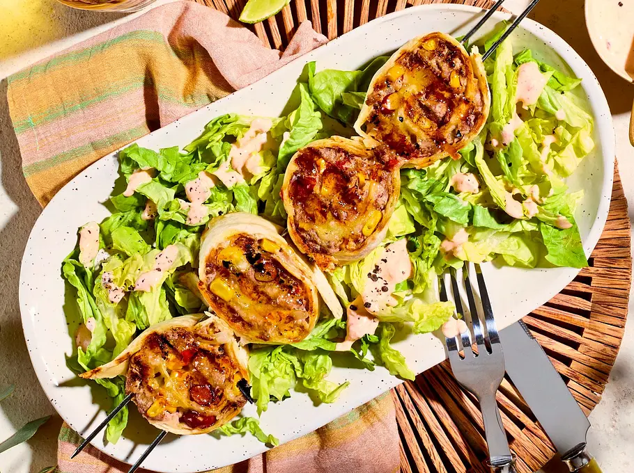 Knusprige Burrito-Taler auf einem Bett aus grünem Salat, angerichtet auf einem ovalen, weißen Teller mit Dressing und Limettenstücken, dazu Holzbesteck.
