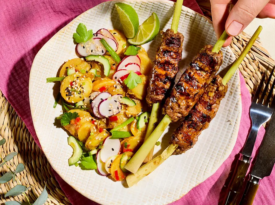 Gegrillte Asia-Hackspieße auf einem weißen Teller mit buntem Kartoffelsalat, Radieschen und Limettenspalten, angerichtet auf einer geflochtenen Unterlage mit pinker Serviette.