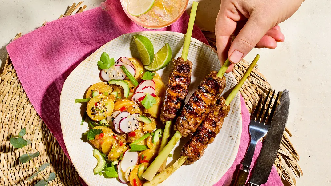 Gegrillte Asia-Hackspieße auf einem weißen Teller mit buntem Kartoffelsalat, Radieschen und Limettenspalten, angerichtet auf einer geflochtenen Unterlage mit pinker Serviette. - Foto: StockFood Studios / Meike Bergmann