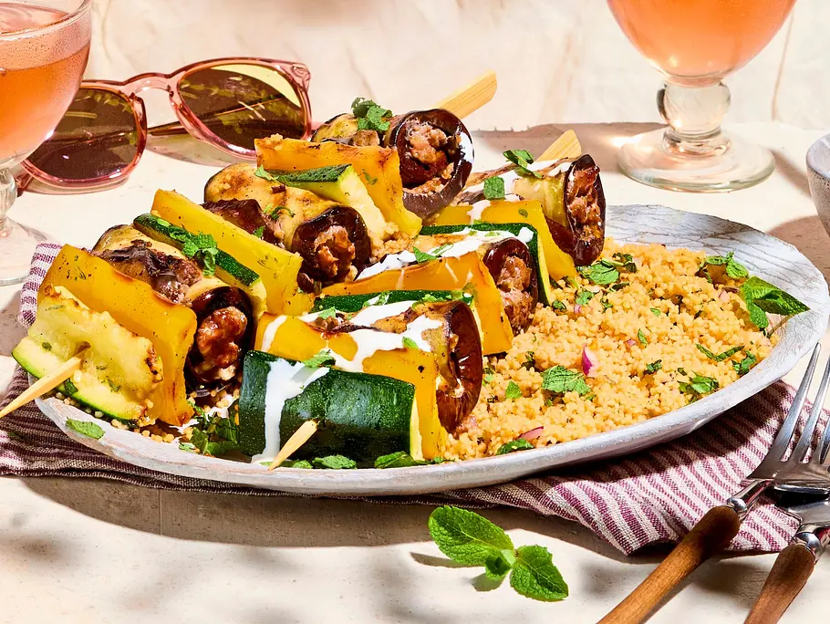 Bunte Ratatouille-Gemüsespieße mit Zucchini und Paprika, serviert auf lockerem Couscous auf einem weißen Teller mit Holzbesteck und gestreifter Serviette.