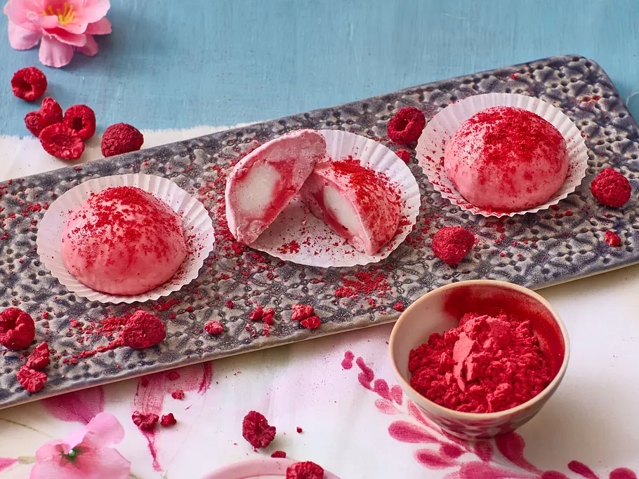 Mochi-Eis mit Himbeersorbet selber machen Drei rosa Mochi-Eiskugeln mit Himbeersorbet, eine angeschnitten, bestäubt mit Himbeerpulver, serviert auf einer rechteckigen Keramikplatte mit weißen Papierförmchen, dekoriert mit gefriergetrockneten Himbeeren.