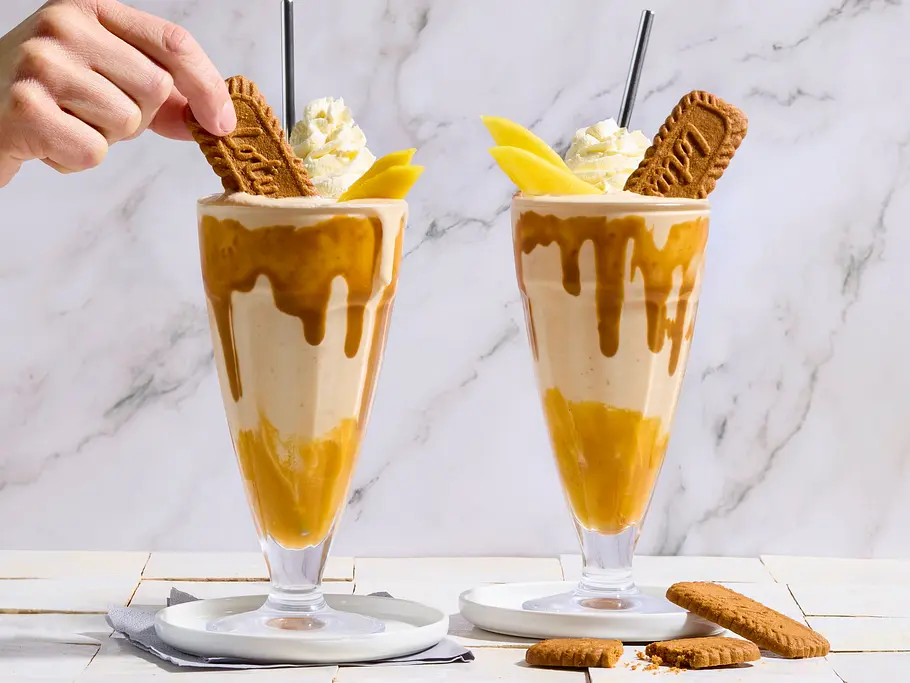 Lotus-Kokos-Milchshake mit Mangosoße Zwei hohe Gläser mit Lotus-Kokos-Milchshake und Mangosoße, garniert mit Sahne, Mangoscheiben und Lotus-Keks, auf weißen Untersetzern, daneben Keksstücke auf gefliester Unterlage.