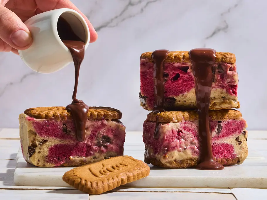 Lotus-Nicecream-Sandwiches mit Kiba-Swirl Drei Lotus-Nicecream-Sandwiches mit Kiba-Swirl auf einem rechteckigen weißen Brett, übergossen mit flüssiger Schokolade, daneben ein einzelner Lotus-Keks und eine Hand mit einer kleinen weißen Kanne.