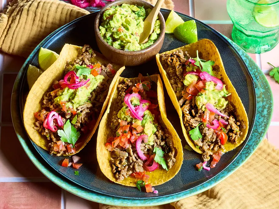 Mexikanische Tacos mit Hackfleisch und Guacamole Rezept Drei knusprige Tacos mit würzigem Hackfleisch, Guacamole, roten Zwiebeln, Tomaten und frischen Kräutern auf einem türkisfarbenen Teller, dazu Limettenspalten, gelbe Stoffserviette sowie kleine Schälchen mit Guacamole, Zwiebeln und Salsa.