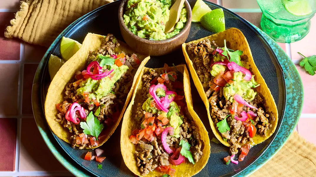 Drei knusprige Tacos mit würzigem Hackfleisch, Guacamole, roten Zwiebeln, Tomaten und frischen Kräutern auf einem türkisfarbenen Teller, dazu Limettenspalten, gelbe Stoffserviette sowie kleine Schälchen mit Guacamole, Zwiebeln und Salsa. - Foto: StockFood Studios / Meike Bergmann