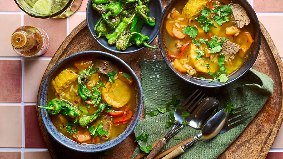 Zwei dunkle Schalen mit chilenischer Cazuela, einem bunten Eintopf mit Rindfleisch, Maiskolben, Kartoffeln, Karotten und Paprika, garniert mit frischem Koriander, serviert auf einem runden Holztablett mit grünem Stoffserviette, Besteck und einer Schale mit gebratenen grünen Paprikaschoten. - Foto: StockFood Studios / Meike Bergmann