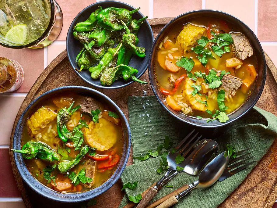 Chilenische Cazuela – traditioneller Eintopf mit Rindfleisch Rezept Zwei dunkle Schalen mit chilenischer Cazuela, einem bunten Eintopf mit Rindfleisch, Maiskolben, Kartoffeln, Karotten und Paprika, garniert mit frischem Koriander, serviert auf einem runden Holztablett mit grünem Stoffserviette, Besteck und einer Schale mit gebratenen grünen Paprikaschoten.