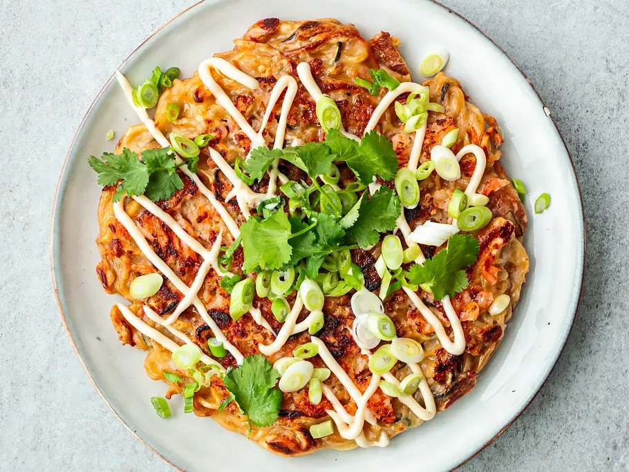 Gebratene Nudelpancake Okonomiyaki-Style, garniert mit Mayonnaise, Frühlingszwiebeln und Koriander, auf einem schlichten weißen Teller serviert.