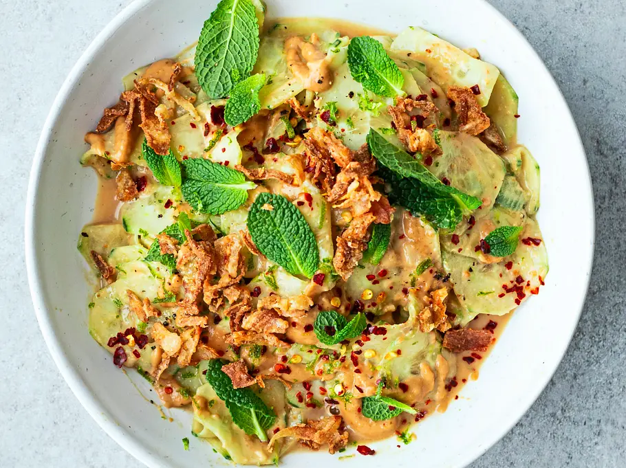 Gurkensalat mit cremigem Erdnuss-Dressing, Chiliflocken, frischer Minze und knusprigen Röstzwiebeln auf einem weißen Teller.