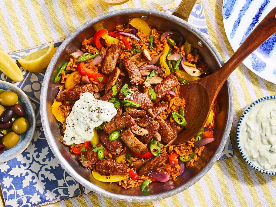 Bunte Reispfanne mit Würstchen-Gyros, serviert in einer Pfanne auf einem gelb-weiß gestreiften Tischtuch, dazu Schälchen mit Oliven, Zaziki und Zitronenspalten.