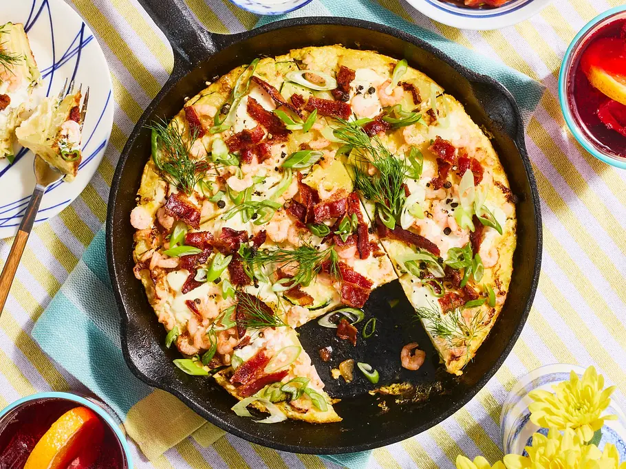 Herzhafte Tortilla mit Nordseekrabben und knusprigem Bacon in einer Eisenpfanne auf gestreiftem Tischtuch, garniert mit frischen Kräutern, dazu Dips und ein Glas mit Getränk und Orangenscheibe.