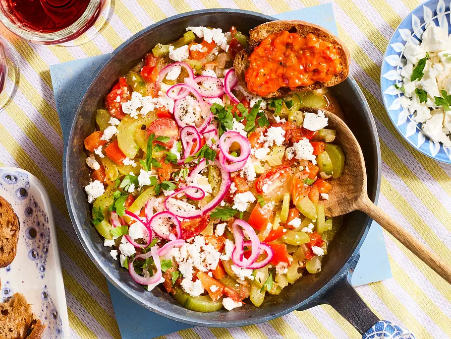 Schmorgurken-Pfanne „Schopska-Style” Schmorgurken-Schopska-Pfanne mit Paprika, Tomaten, Zwiebeln und zerbröseltem Feta, serviert in einer Pfanne auf gelb-weiß gestreiftem Tischtuch, dazu geröstetes Brot, Ajvar und zwei Gläser rotes Getränk.
