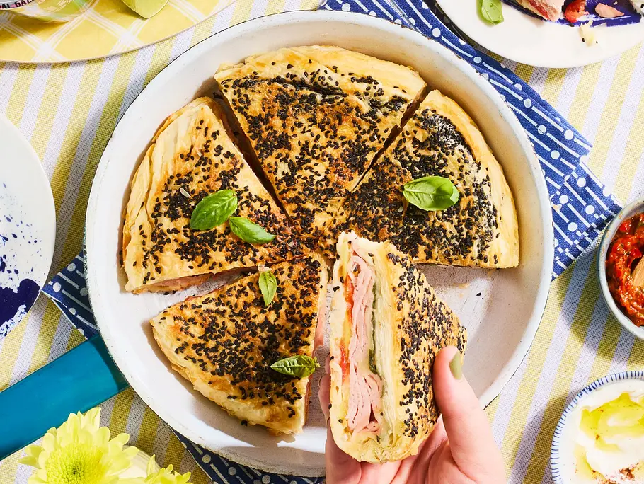 Pfannen-Burek mit Leberkäse, Tomaten und Käse im knusprigen Sesammantel, serviert in Tortenstücken auf einem weißen Teller, dazu Schälchen mit Dip und gelb-weiß gestreiftes Tischtuch.