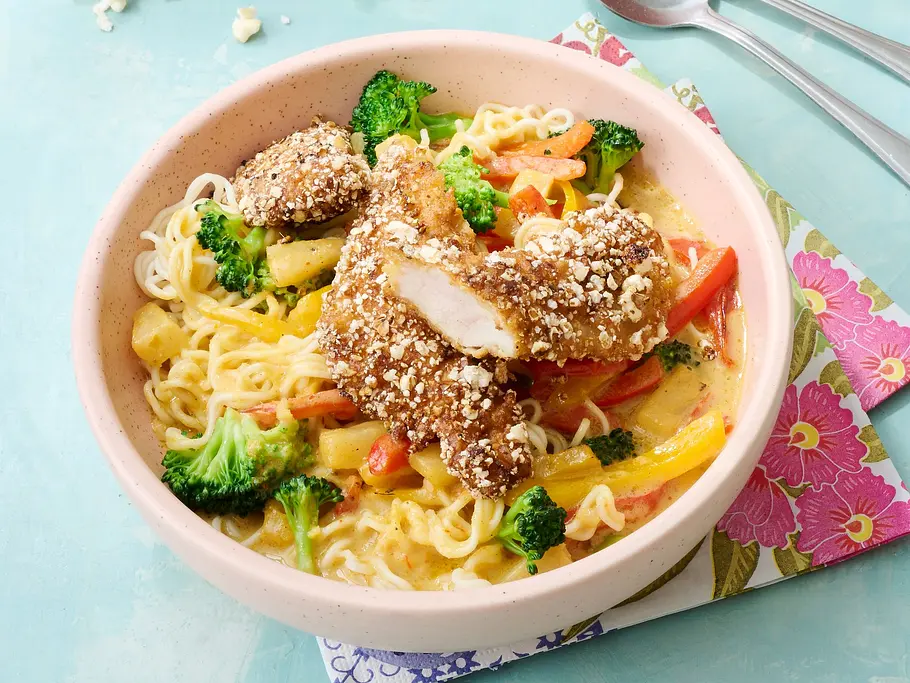 Popcorn-Schnitzel Knusprige, mit Popcorn panierte Mini-Schnitzel auf einer bunten Gemüse-Curry-Nudelpfanne mit Brokkoli, Paprika und Möhren, angerichtet in einer rosa Keramikschale auf pastellfarbener Unterlage.