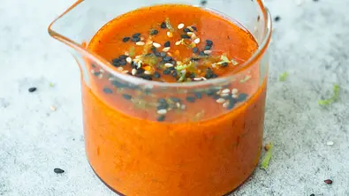 Feurig-rotes Honig-Senf-Dressing mit Hot Honey und Harissa in einem Glasbecher, bestreut mit schwarzem und weißem Sesam, auf heller Steinplatte serviert. - Foto: Simone Neufing/MOE‘S QUICK & EASY FOOD 