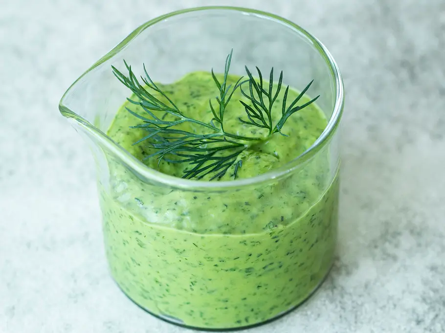 Cremiges, grünliches Honig-Senf-Dressing mit Kräutern und Avocado in einem Glasbecher, garniert mit frischem Dill auf heller Steinplatte.