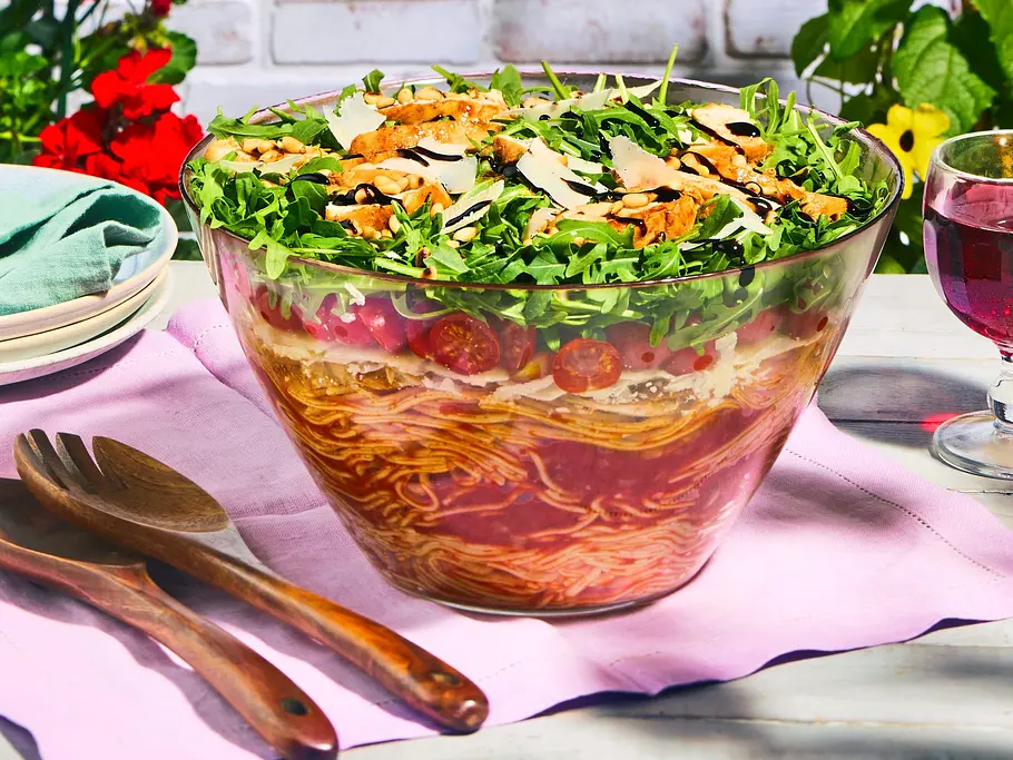 Italienischer Spaghetti-Schichtsalat mit Hähnchen Große Glasschüssel mit geschichtetem Spaghetti-Salat, oben mit Rucola, Tomaten, gehobeltem Parmesan und Hähnchenstückchen, auf einem hellen Holztisch mit rosa Serviette und Holz-Besteck.