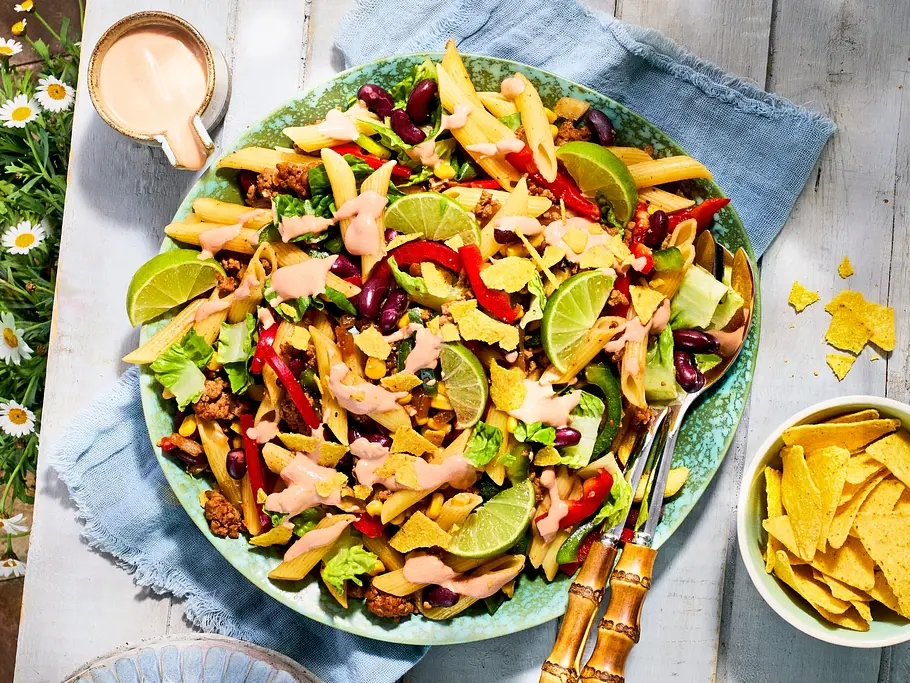 Großer grüner Teller mit buntem Nudelsalat, verfeinert mit Kidneybohnen, Paprika, Limettenstücken, Salat, zerbröselten Tortilla-Chips und einem rosa Dressing, daneben eine Schale mit weiteren Tortilla-Chips auf hellem Holztisch.