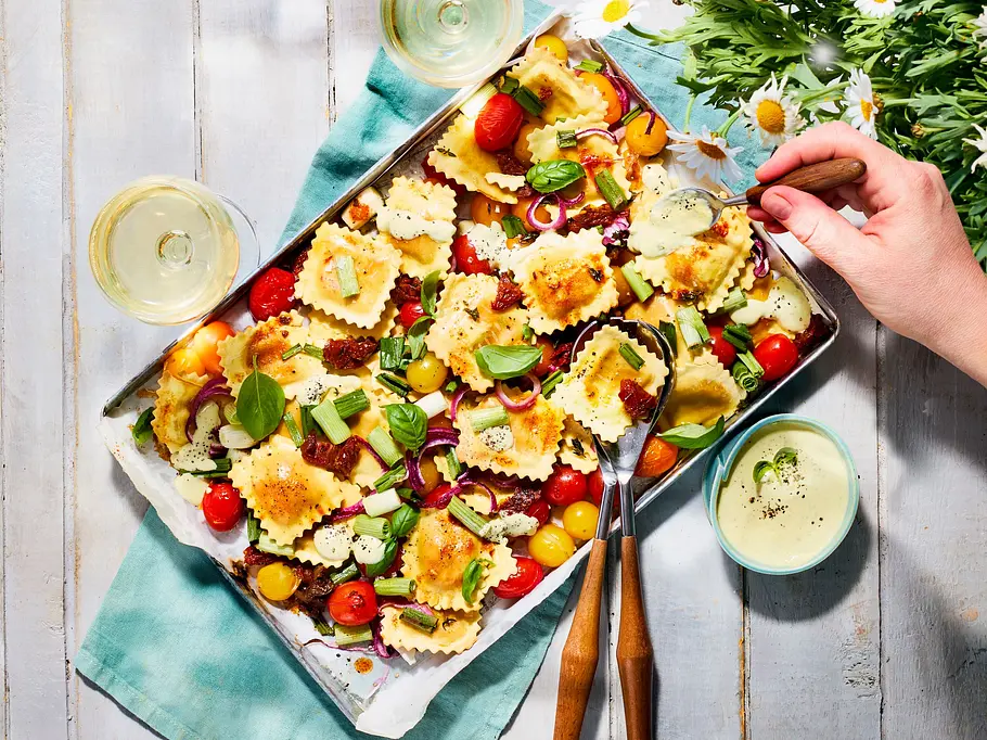 Lauwarmer Ravioli-Salat vom Blech Blech mit buntem Ravioli-Salat, garniert mit frischen Tomaten, roten Zwiebeln, Basilikum und etwas Käse auf einem weißen Holztisch; daneben liegen ein türkises Tuch und ein Besteck.